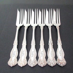 May include: A set of six silver-toned metal cocktail forks arranged in a row. Each fork features three tines, with the left tine being wider and pointed. The handles are decorated with embossed floral patterns and a central engraved monogram letter.