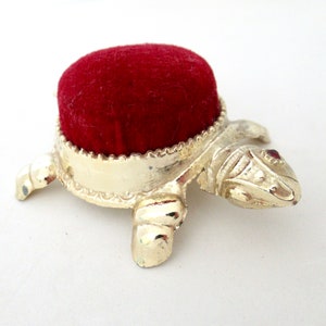 May include: A gold turtle pincushion with a red velvet top. The turtle has a detailed design and a red jewel on its head.