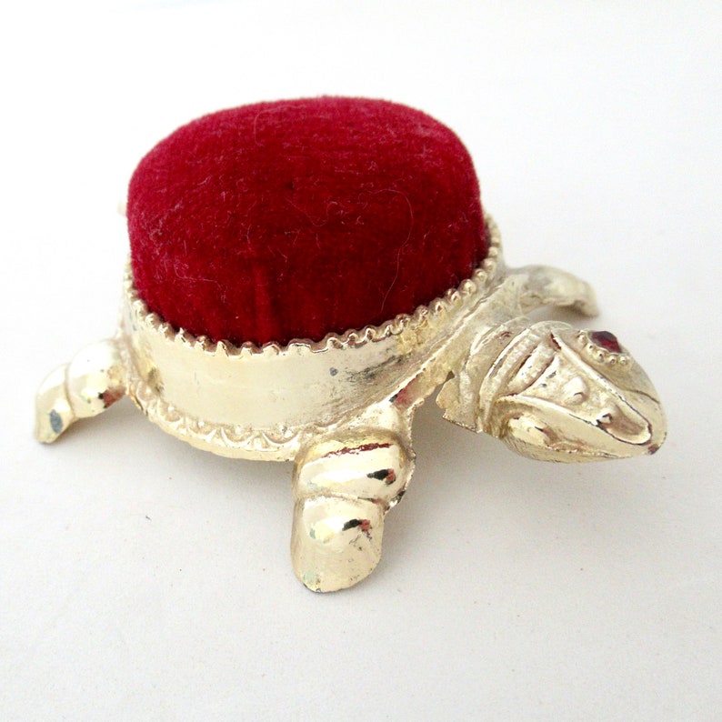 May include: A gold turtle pincushion with a red velvet top. The turtle has a detailed design and a red jewel on its head.