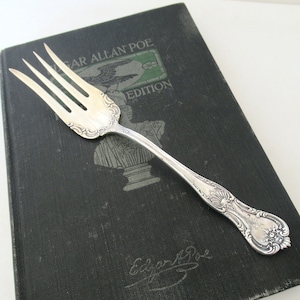 May include: A silver fork with an ornate design, resting on a black book with the text "Edgar Allan Poe Edition".