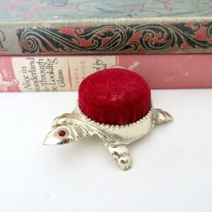 May include: A silver turtle pincushion with a red velvet top. The turtle has red jewel accents on its head.