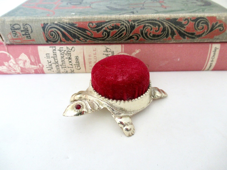 May include: A silver turtle pincushion with a red velvet top. The turtle has red jewel accents on its head.