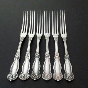 May include: A set of six silver-toned metal forks arranged in a row. Each fork features a four-tine head and a handle decorated with embossed floral patterns at the base and neck, with a smooth, plain central section on the handle.