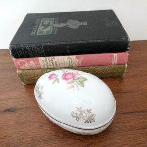 May include: White oval trinket box with a floral design featuring pink roses and gold accents. The box is closed and sitting on a wooden surface.