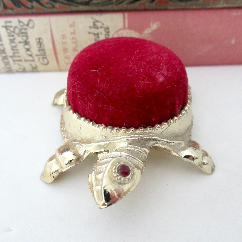 May include: A small, gold-colored turtle pincushion with a red velvet top. The turtle has a red jewel on its shell.