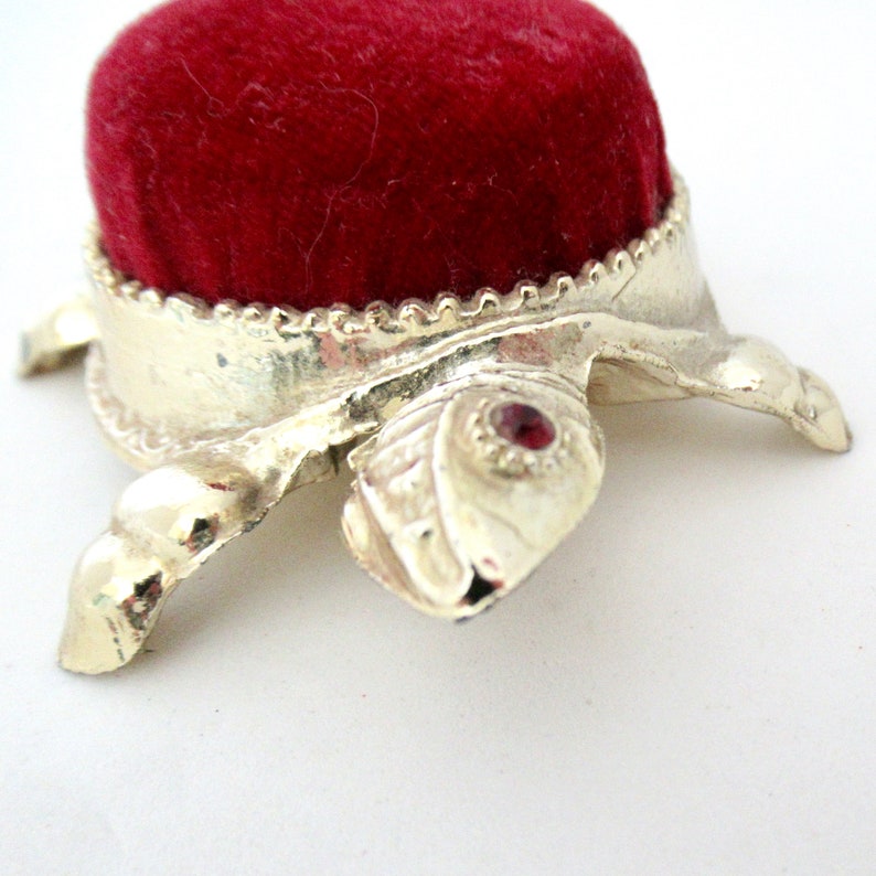 May include: A small, red velvet pincushion sits atop a gold turtle-shaped base. The turtle has a red jewel in its eye.