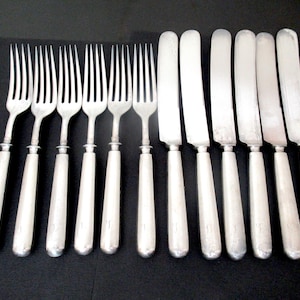 May include: A set of twelve silver-toned flatware pieces consisting of six forks and six knives. Each piece features a smooth, cylindrical handle and a polished metal finish. The forks have four tines, while the knives have a rounded blade.
