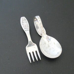 May include: A vintage silver fork and spoon set for a child. The fork has a detailed embossed design of a bear and the words "Baby's Fork" and "Sterling". The spoon has the words "Toot Toot Riding Hood" on the handle.