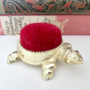 May include: A gold turtle pincushion with a red velvet top. The turtle has a detailed design and is sitting on a white surface.