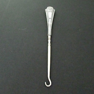 May include: A silver Art Deco style button hook with a decorative handle and a curved hook end. The handle features an ornate design with geometric patterns. The hook is designed to assist with buttoning garments.