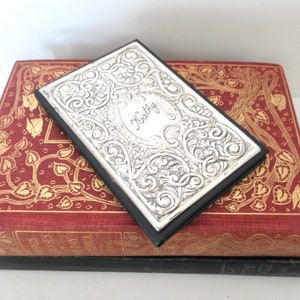 May include: A silver-colored journal with an ornate design and the name "Kathy" engraved on the front. The journal is closed and resting on top of a red and gold book.