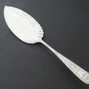 May include: A silver cake server with an ornate handle and a scalloped edge. The handle is decorated with a floral design and a bird.