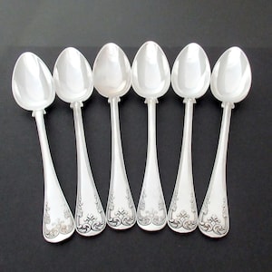 May include: A set of six silver-toned metal spoons arranged in a row. Each spoon features a polished oval bowl and a handle decorated with intricate, embossed scrollwork and floral patterns at the base, showcasing a reflective metallic finish.