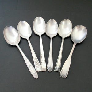 May include: Six vintage silver spoons with oval bowls and ornate handles are arranged on a dark surface. The spoons vary in handle design, with some featuring floral or geometric patterns. The spoons are reflective and show signs of age.