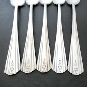 May include: Five silver spoons arranged in a row, each with a fan-shaped handle design. The handles have a series of vertical lines and a decorative floral pattern near the base. The spoons are set against a dark background.