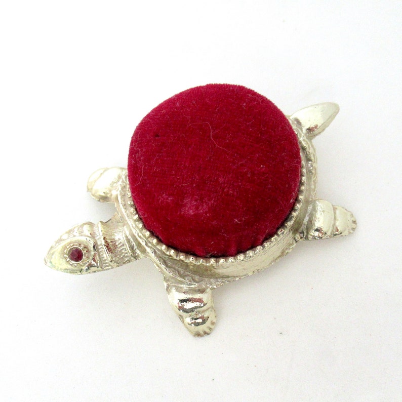 May include: A silver turtle pincushion with a red velvet top. The turtle has a red jewel in its eye.