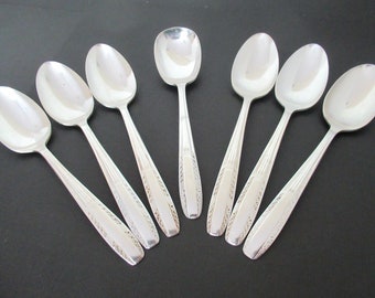 May include: A collection of seven silver spoons arranged on a dark surface. The spoons have oval bowls and decorative handles with etched patterns. The spoons are reflective, showing the surrounding environment.