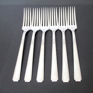 May include: Six silver forks with a geometric design on the handles are arranged on a dark surface. The forks have long, slender tines and a decorative pattern near the top of the handle.