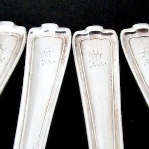 May include: Four silver-toned metal spoon handles arranged side by side. Each handle features a flared, stepped top edge and a central engraved monogram consisting of the letters 'M' and 'H' intertwined in a decorative, gothic-style script.