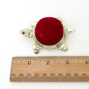 May include: A silver turtle pincushion with a red velvet top. The turtle has a red jewel for an eye. The pincushion is about 1 inch in diameter.