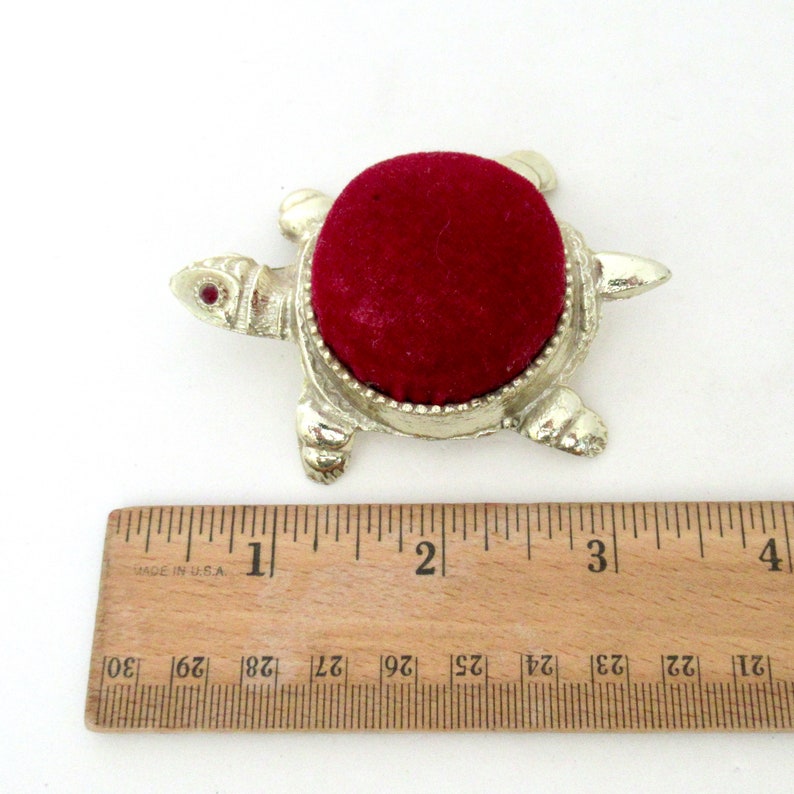 May include: A silver turtle pincushion with a red velvet top. The turtle has a red jewel for an eye. The pincushion is about 1 inch in diameter.
