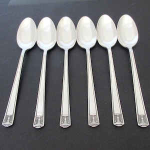 May include: Six silver spoons arranged in a row. Each spoon has an oval bowl and a long, rectangular handle with a decorative design at the end. The spoons are made of a shiny, reflective metal.