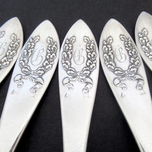 May include: Close-up of five silver spoons. Each spoon features an engraved design of a laurel wreath with a bow and the letter "B" in the center. The spoons are arranged in a fan shape against a dark background.