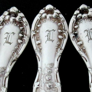 May include: Close-up of several antique silver spoons. Each spoon features an ornate floral design with the letter "I" engraved on the handle. The spoons have a bright, reflective finish against a black background.
