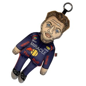 Könnte beinhalten: Ein Plüschpuppen-Schlüsselanhänger mit einer stilisierten Illustration einer Person mit braunen Haaren und blauen Augen. Die Puppe trägt einen dunkelblauen Rennanzug mit roten Akzenten, der mit MIRACLE, WINGS, The Bull, Mobil und einem roten Stier-Logo bedruckt ist.