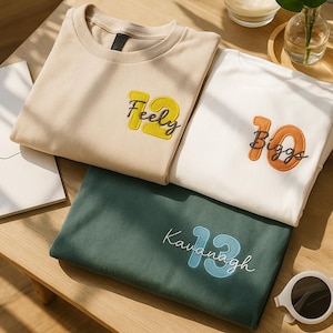 May include: Four folded t-shirts in beige, white, and green are displayed on a wooden surface. Each shirt features a number and a name embroidered on the front. The beige shirt has "Feely 12", the white shirt has "Biggs 10", and the green shirt has "Kavanagh 13".