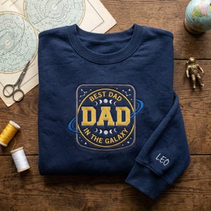 May include: A navy blue sweatshirt featuring an embroidered patch with the text "BEST DAD IN THE GALAXY" in gold and white. The sleeve cuff is embroidered with the name "Leo". The sweatshirt is displayed on a wooden surface with decorative items.