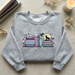 May include: A grey sweatshirt with an embroidered design of books in purple, blue, and green. The design includes a wolf howling at a yellow moon, a butterfly, flowers, stars, and bats. The sweatshirt has a crew neck and long sleeves.