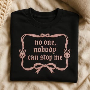 May include: Black sweatshirt with the embroidered text "no one, nobody can stop me" in pink. The design includes a pink frame with bunny heads and bows. The sweatshirt is folded on a beige surface.