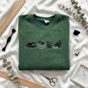 May include: A dark green sweatshirt with a crew neck. It has an embroidered design of four black cats in different poses. One cat is in a box, another is eating from a bowl. The sweatshirt is made of a soft material.