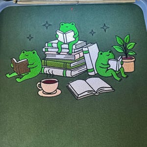 May include: Embroidered design with a dark green background, depicting three green frogs reading books, a cup of coffee, a potted plant, and stacks of books. Four star shapes are also embroidered. The design is likely for clothing or accessories.