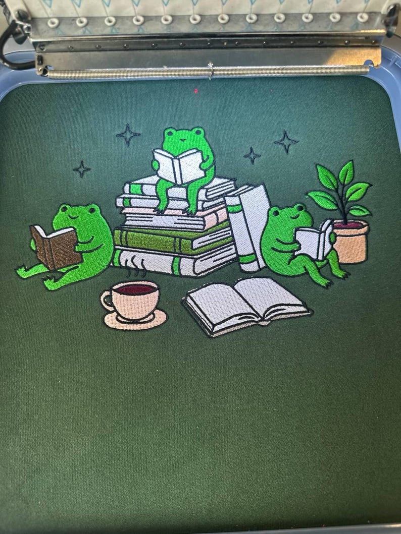 May include: Embroidered design with a dark green background, depicting three green frogs reading books, a cup of coffee, a potted plant, and stacks of books. Four star shapes are also embroidered. The design is likely for clothing or accessories.