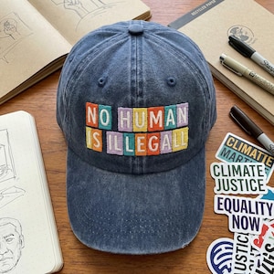 May include: A dark blue denim baseball cap featuring the embroidered phrase "NO HUMAN IS ILLEGAL" in multi-colored block letters. The cap is on a wooden surface with notebooks and stickers.