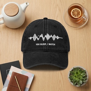 May include: Black denim baseball cap with a white sound wave graphic and the text "YOU SLEEP, I WATCH". The cap is on a wooden surface with a teapot, a cup of tea, a succulent plant, and notebooks.