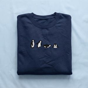 May include: A navy blue sweatshirt with an embroidered design of four penguins in different poses. The penguins are black and white with orange detailing. The sweatshirt is folded on a light blue surface.
