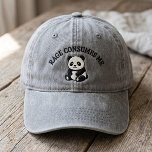 May include: A grey baseball cap with a distressed fabric finish. The front features an embroidered black and white panda bear sitting in the center, with the black embroidered text "RAGE CONSUMES ME" arched in a semi-circle above the panda.