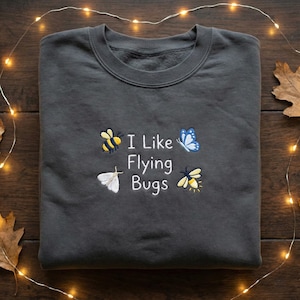 May include: Dark grey sweatshirt featuring embroidered insects and the text "I Like Flying Bugs." The design includes a bee, butterfly, moth, and firefly. The sweatshirt is folded on a wooden surface, with fairy lights and leaves in the background.