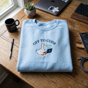 Peut inclure: Un sweat-shirt bleu clair avec la phrase brodée "OFF TO CODE" et un canard de dessin animé utilisant un ordinateur portable. Une tasse noire avec "Hello World" est à gauche. Un ordinateur portable, un téléphone et un clavier sont en arrière-plan.