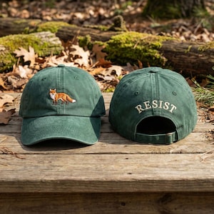 Könnte beinhalten: Zwei grüne Baseballkappen. Eine Kappe zeigt einen gestickten Fuchs, die andere das Wort „RESIST“ auf der Rückseite. Beide Kappen haben einen verstellbaren Riemen und einen gebogenen Schirm. Die Kappen werden auf einer Holzoberfläche präsentiert.