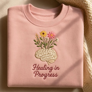 Peut inclure: Sweat-shirt rose clair avec une broderie représentant un cerveau d'où sortent des fleurs. La broderie comprend le texte "Healing in Progress". Les fleurs sont jaunes, roses et blanches. Col rond.