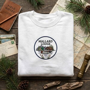 May include: White sweatshirt featuring a circular embroidered patch with the words "Mallard Lodge." The patch depicts a duck on water, mountains, and a cabin. The sweatshirt is surrounded by outdoor-themed items.