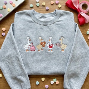 May include: A gray sweatshirt featuring an embroidered design of four geese wearing chef hats. Each goose is holding a different baked good: a cake, bread, a bowl, and a tray of pastries. The sweatshirt is surrounded by heart-shaped candies.
