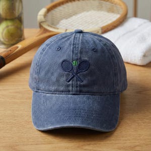 May include: A blue denim baseball cap featuring an embroidered tennis racket and ball design. The cap has a curved peak and a button on top. Tennis equipment is visible in the background.