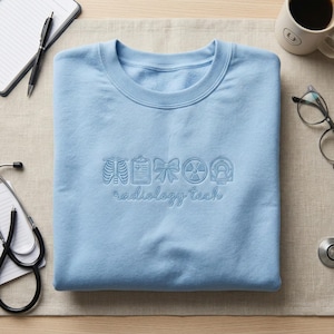 May include: A pale blue sweatshirt featuring embroidered radiology symbols and the text "radiology tech". The design includes a rib cage, clipboard, bow, radiation symbol, and a medical device. The sweatshirt is neatly folded on a neutral surface.