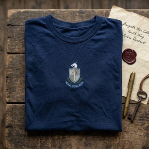 May include: A navy blue t-shirt with a crew neck, displaying an embroidered crest with a dragon, cross, and the words "War College." The shirt is folded on a wooden surface, with a vintage paper and accessories.