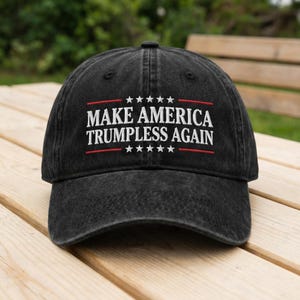 May include: A black, distressed baseball cap with the text "MAKE AMERICA TRUMPLESS AGAIN" in white, with red accents. The hat has a curved brim and a fabric strap closure. The cap is sitting on a wooden surface.